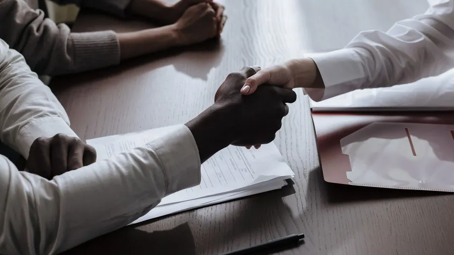 Two people shaking hands over signed contract documents at a meeting table, closing an equity and hiring agreement for a startup marketing leader