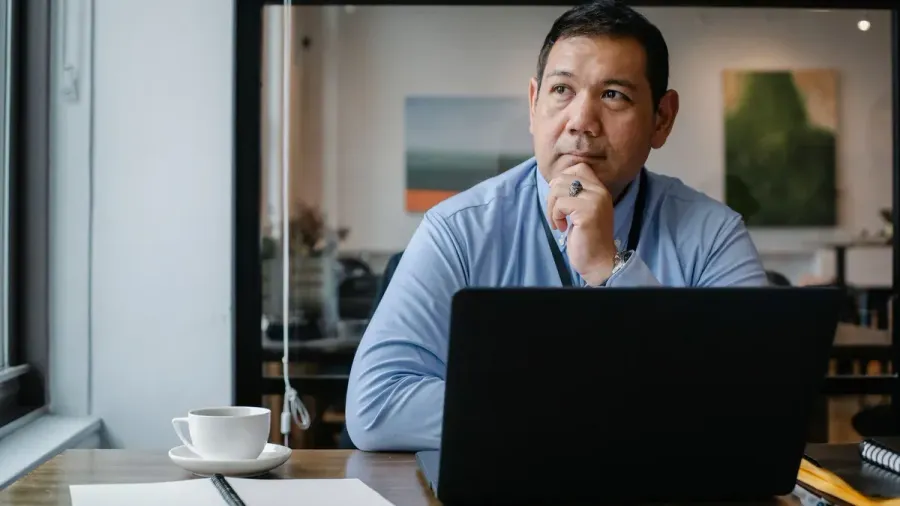 A small-business owner sits at a clean desk with a laptop, coffee and a blank notepad, pausing thoughtfully to weigh a decision — choosing the outcome, not the menu of options