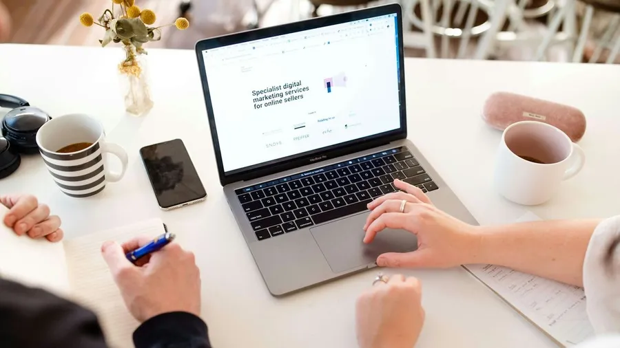 Two people reviewing a digital marketing services website on a laptop over coffee, a notepad and pen on the table — weighing what it costs to outsource marketing