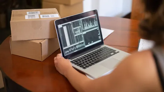 A small business owner reviewing sales charts on a laptop at a table surrounded by shipping boxes — figuring out what actually works in small business marketing