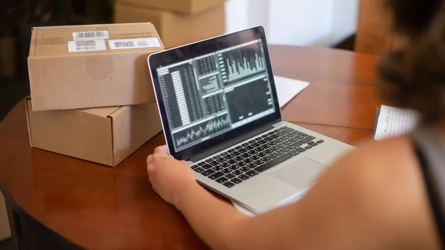 A small business owner reviewing sales charts on a laptop at a table surrounded by shipping boxes — what works in small business marketing in 2026